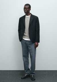 Blazer in tessuto nero, maglione in maglia grigio chiaro, camicia bianca, jeans blu e mocassini neri. Texture lisce con design e vestibilità su misura.