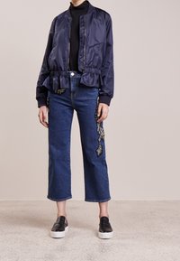 Chaqueta bomber de satén azul marino con dobladillo fruncido, combinada con jeans cortos azules que presentan bordados florales metálicos en los laterales y zapatos negros tipo slip-on.