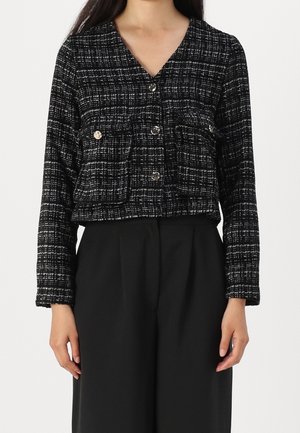 Kvinde iført en sort- og hvide ternet knapjakke med forlommer og sorte bukser med vide ben, stående foran en enkel baggrund.