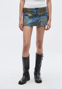 Τζιν μίνι φούστα με χρυσά και ασημένια τρουκς, με εμπρόσθιο κλείσιμο με κουμπιά και εφαρμοστή γραμμή. Συνοδεύεται από μαύρες δερμάτινες μπότες μέχρι το γόνατο.