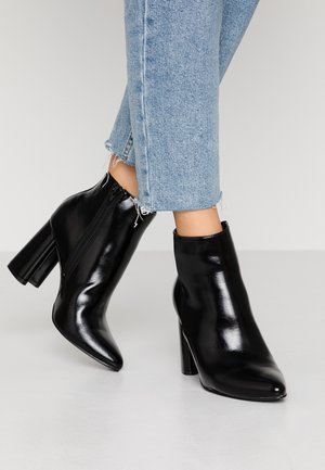 Skinnende sorte læderankelstøvler med blokhæle båret med lyseblå opkrogede jeans mod en ensfarvet baggrund.