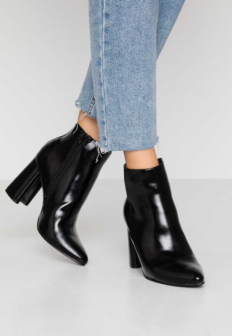 Bottes noires en cuir brillant à talons carrés portées avec un jean court bleu clair sur un fond uni.