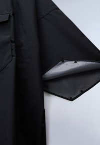 Sort ærme med trykknapper delvist åbnet, der afslører en grå inderside og teksturerede stofdetaljer på beklædningsgenstanden.