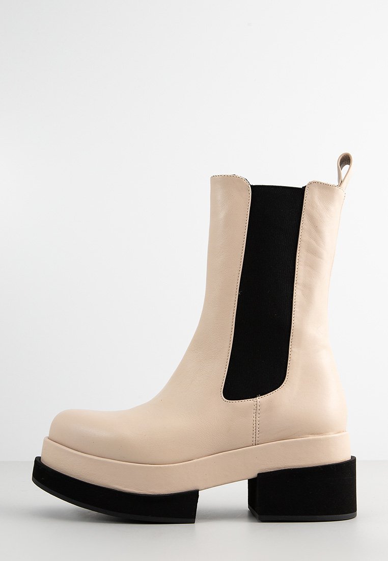 Bota de cuero beige hasta el tobillo con laterales elásticos negros, puntera redonda, tacón de plataforma negro grueso y una lengüeta en la parte posterior. Textura suave.