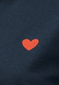 Μικρή κόκκινη κεντητή καρδιά σε σκούρο μπλε ύφασμα, τοποθετημένη στο κέντρο του κάδρου, δείχνοντας λεπτομέρειες υφής και ραφής.