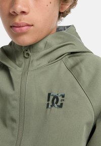 Vihreä huppari, jossa on teksturoitu pinta. Etuosassa on kohotettu musta DC-logo rinnassa ja edessä vetoketjukiinnitys.