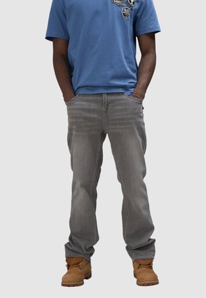 Grijze denim jeans met een rechte pasvorm, blauwe shirt met korte mouwen met patches, en bruine enkelboots met veters.