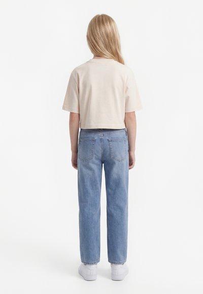 Blondynka stojąca tyłem, ubrana w skrócony beżowy t-shirt, jasnoniebieskie dżinsy i białe trampki na białym tle.
