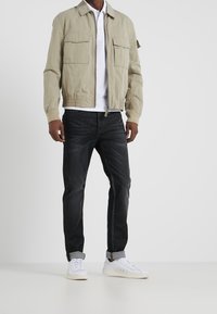 Chaqueta bomber beige claro con dos bolsillos frontales, puños elásticos y cinturilla. Combinada con jeans de mezclilla oscuros y zapatillas blancas.