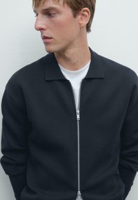 Junger Mann in einem schwarzen Reißverschluss-Pullover über einem weißen Hemd, der zur Seite schaut, vor einem schlichten hellen Hintergrund.