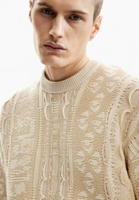 Közeli kép egy fiatal férfiról, aki texturált, krémszínű kötött pulóvert visel aprólékos mintákkal, és kis fülbevaló van a bal fülében.