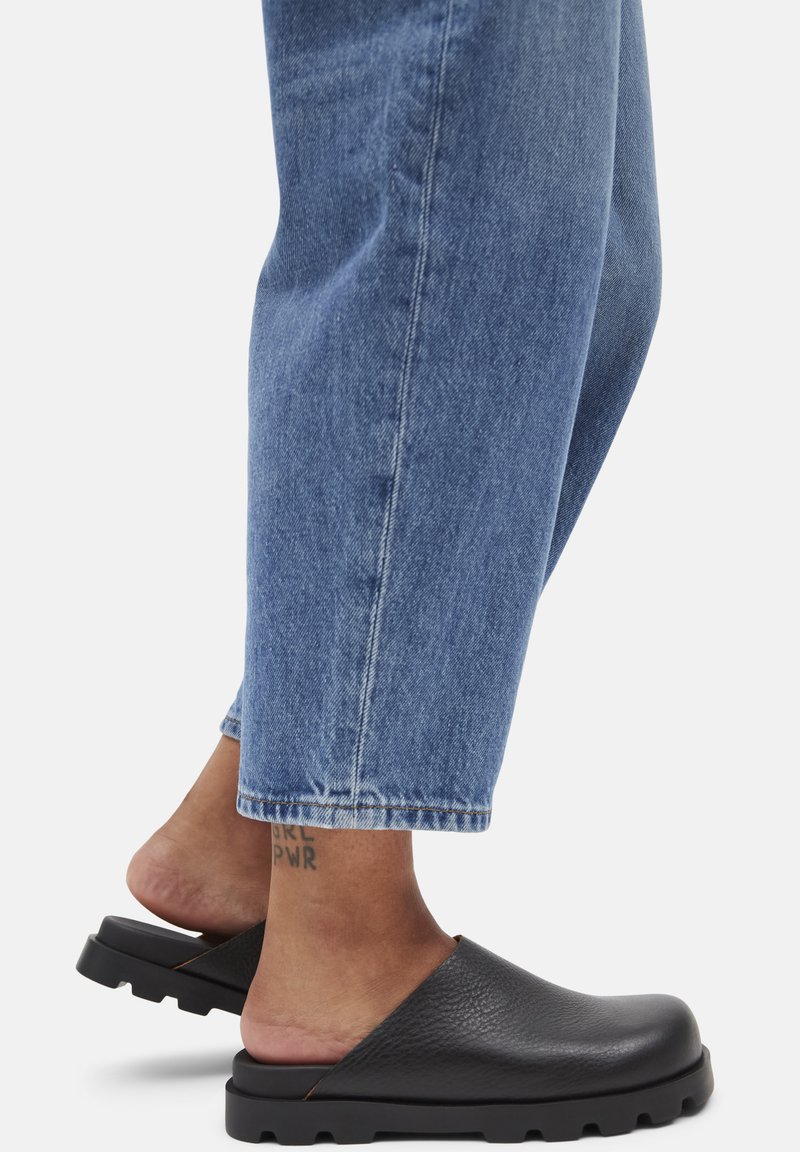Sabots en cuir noir avec une finition texturée et une épaisse semelle en caoutchouc. Associés à un jean en denim bleu clair, avec une coupe courte.