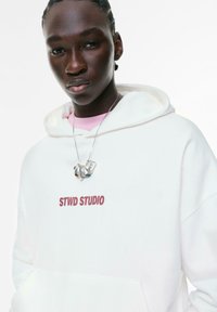 Ung person med fletninger iført en hvid hoodie med teksten "STWD STUDIO" og et sølv chunky halskæde foran en hvid baggrund.