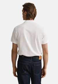 Weißes kurzärmeliges Poloshirt von hinten gesehen, kombiniert mit dunklen Denim-Jeans und einem schwarzen Gürtel mit braunem Lederakzent.