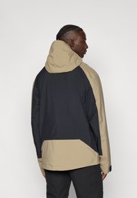 Μπουφάν με κουκούλα, με σχέδιο σε μπλοκ χρωμάτων σε μπεζ και σκούρο ναυτικό μπλε. Κατασκευασμένο από ελαφρύ ύφασμα με ομαλή υφή και ελάχιστα μεταλλικά εξαρτήματα.