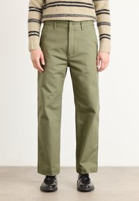 Green cotton pants with a straight-leg cut, featuring a button closure and side pockets, paired with black shoes. Textured sweater visible above.