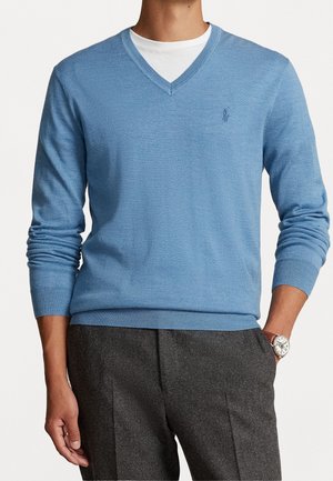 Man draagt een lichtblauwe V-hals trui over een wit T-shirt, donkergrijze broek en een horloge met een bruine leren band.