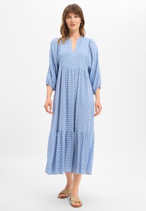 Woman with shoulder-length hair wearing a light blue patterned long dress and beige sandals, standing against a white background.