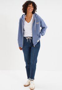 Felpa azzurra, misto cotone, con tasche frontali e cordoncino. Indossata sopra una t-shirt bianca a collo a V e abbinata a jeans blu scuro.