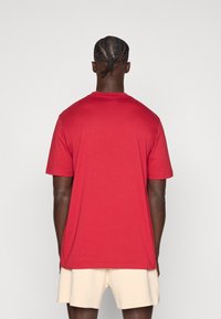 Sarkana kokvilnas T-krekls ar īsām piedurknēm un apaļu kakla izgriezumu, ar gludu tekstūru un brīvu piegriezumu. Aizmugures skats rāda pastiprinātas šuves.