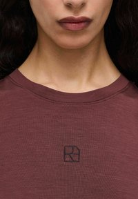 Bordeauxfarvet bomulds t-shirt med rund halsudskæring og et lille sort broderet logo på brystet. Glat, tekstureret stof med subtile linjer.