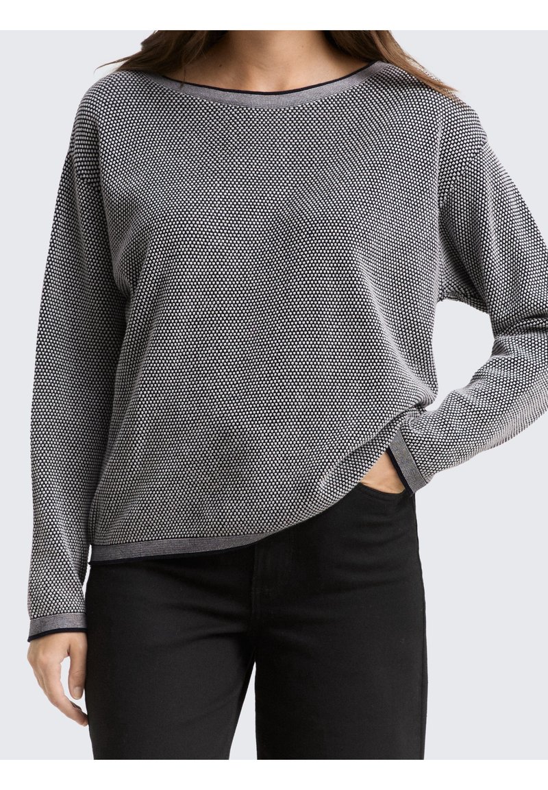 Femme portant un pull à manches longues noir et blanc à motifs avec un pantalon noir, une main dans la poche, fond neutre.