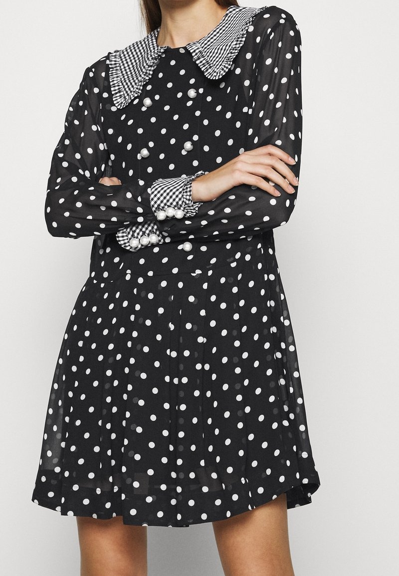 Robe noire à pois blancs ; elle présente un grand col à carreaux et des détails à boutons, des manches longues avec des poignets à pois, et une jupe évasée.