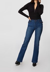 Černý dlouhý rukáv s límcem na zip, spárovaný s tmavě modrými rozšířenými džínami s dekoračními doplňky v oblasti pasu a hladkou texturou.