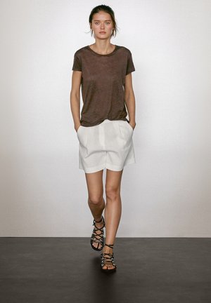 T-shirt in lino marrone, pantaloni corti plissettati bianchi e sandali neri con cinturini. La modella ha un'acconciatura naturale e si trova davanti a uno sfondo neutro.