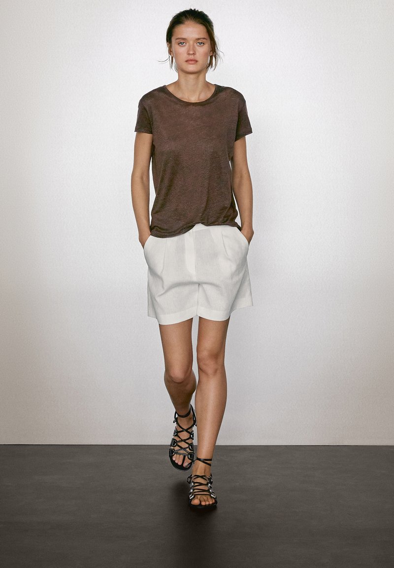 Brun linned t-shirt, hvide plisserede shorts og sorte strop sandaler. Modellen har en naturlig frisure og står foran en neutral baggrund.
