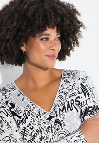 Top a maniche corte in tessuto bianco con motivi in testo scritto a mano neri. Design a V con una texture liscia e una leggera lucentezza.