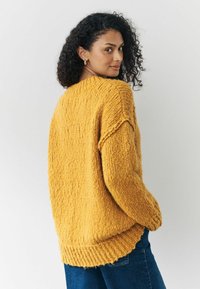 Struktureret, sennepsgul strikket sweater med en bred ribbet kant og sænkede skuldre, parret med blå jeans i en afslappet indstilling.