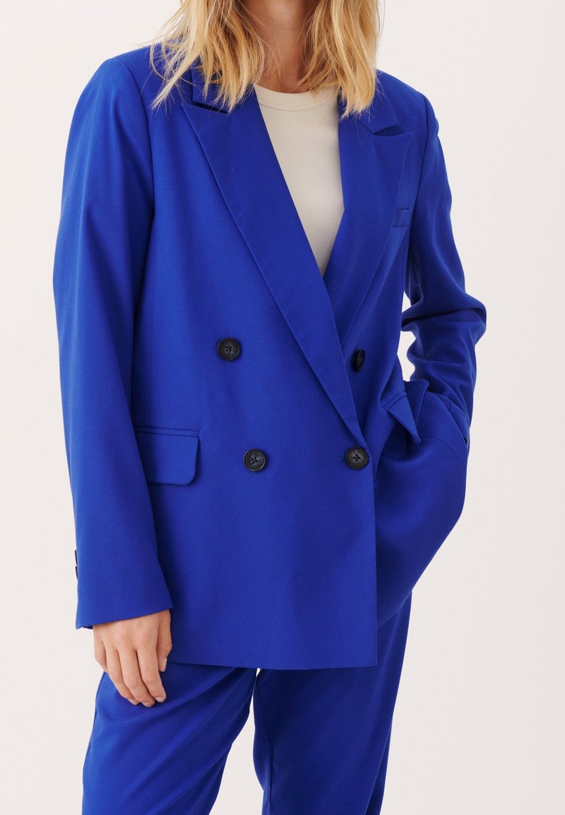 Blazer bleu à double boutonnage avec revers crantés, boutons noirs, deux poches avant et épaules structurées ; tissu lisse avec une coupe ajustée.