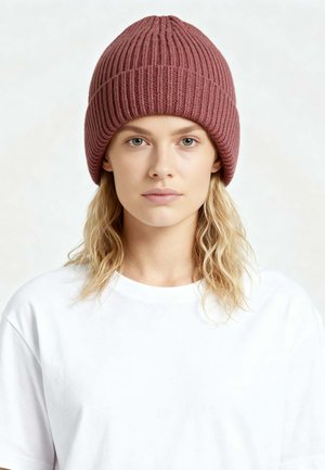 Giovane donna con capelli biondi ondulati indossa un berretto lavorato a maglia rosa polvere a coste e una maglietta bianca semplice, rivolta in avanti con espressione neutra.