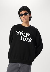 Čierna mikina s bielym nápisom "New York", okrúhlym výstrihom, zníženými ramenami a rebrovaným lemom; nosená s čiernymi slnečnými okuliarmi a sivými nohavicami.