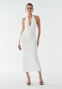 Robe blanche à encolure halter avec un décolleté en V profond et un devant froncé. Silhouette moulante et extensible, atteignant le mollet. Talons à bride de cheville noirs.