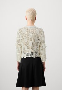 Kardigan z dzianiny koronkowej w kremowym kolorze z rękawami do łokcia, ozdobiony misternym kwiatowym wzorem i falistym dołem, noszony na czarnej sukience.