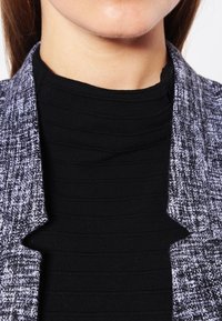 Close-up van de nek en onderkant van het gezicht van een persoon die een zwarte ribbeltrui met hoge hals en een grijze tweed jas draagt.