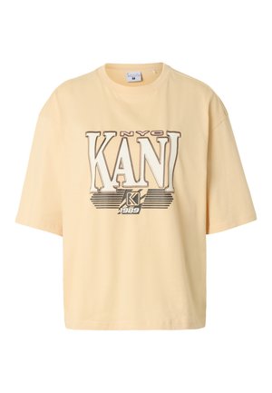 Ung kvinde med blondt hår har en lys beige oversized T-shirt med "NYC KANI 1989" logo og mørkebrune bukser mod en enkel baggrund.