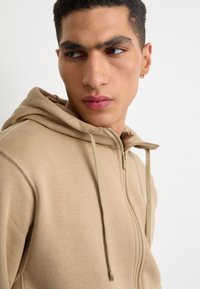 Sweatshirt bege com capuz, fecho éclair completo e cordão. Textura de tecido suave e detalhes de costura visíveis. Sem logótipos ou padrões.