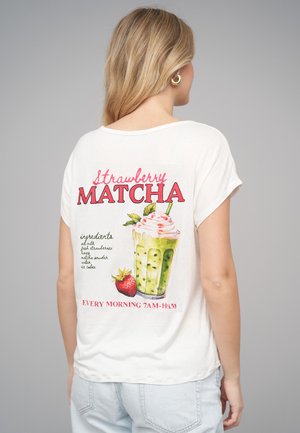 Pitkähiuksinen vaalea nainen, joka käyttää valkoista T-paitaa, jossa on "Strawberry Matcha" -juoman kuvitus ja ainesosaluettelo takana.