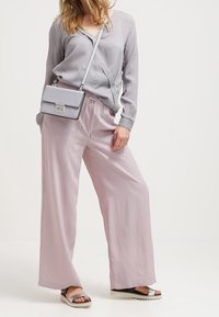 Světle šedá halenka s dlouhými rukávy a výstřihem do V, doplněná bledě růžovými širokými kalhotami. Oblečení zahrnuje černé sandály se stříbrnými detaily.