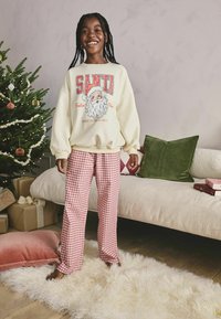 Kermainen collegepaita, jossa on punainen "SANTA" -grafiikka ja joulupukin kuvasto. Punavalkoiset ruudulliset pyjamahousut, rentoa istuvuutta.