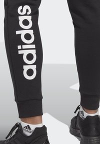 Fekete pulóvernadrág fehér "adidas" logóval, amely függőlegesen fut a bal lábszáron. A nadrág szára szűkített, fekete sportcipővel és zoknival párosítva.