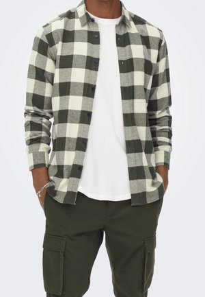 Man draagt een groen-wit geruit blousehemd met knopen over een effen wit T-shirt en donkergroene cargobroek, handen in de zakken.