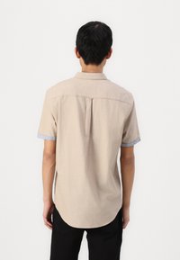 Camisa de manga corta en tejido beige claro con cuello abotonado, con detalles azul en las mangas y un dobladillo redondeado en la parte posterior.