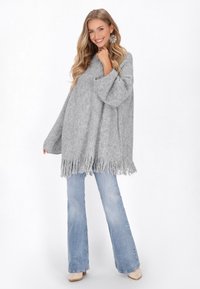 Grauer gestrickter Poncho mit fransigem Saum, lockere Passform, lange Ärmel, kombiniert mit hellblauen Schlagjeans und beigen Schuhen.