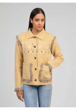 Femme portant une veste beige boutonnée avec des motifs brodés et des détails à franges sur les poches, les manches et les épaules, associée à un jean bleu clair.