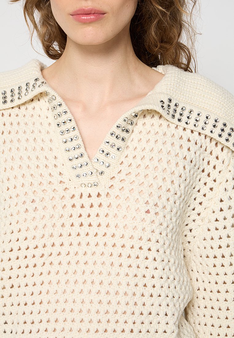 Cremfarbener Strickpullover mit V-Ausschnitt, der metallische Nieten entlang des Kragens und eine lockere, offene Webstruktur aufweist.