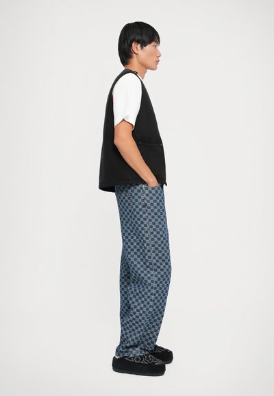 Un vest negru fără mâneci peste un tricou alb, asociat cu pantaloni albaștri cu un model. Detalii notabile includ accentele de buzunare și încălțăminte închise la culoare.